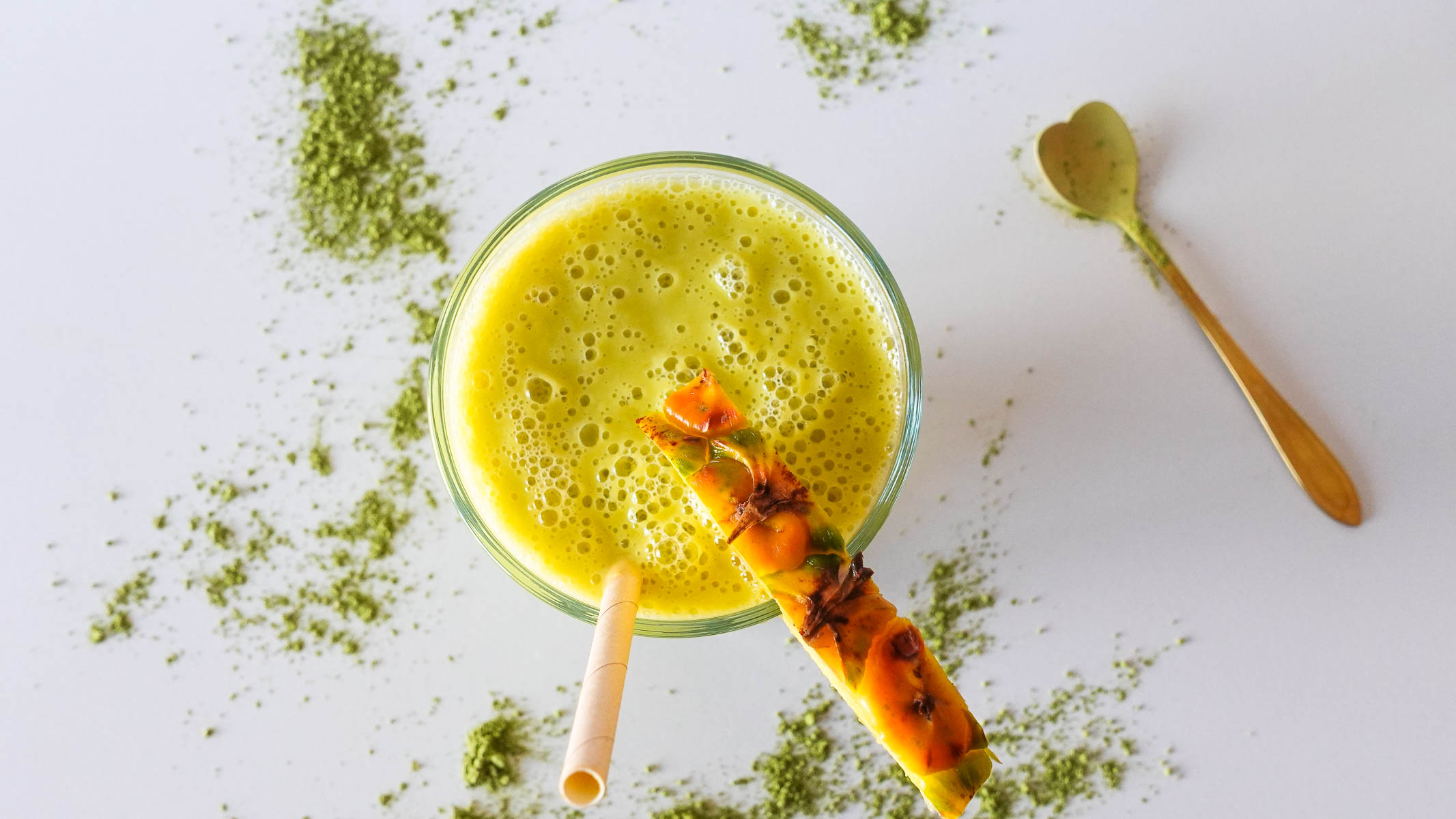 Matcha Mango Pineapple Smoothie in a glass with matcha powder on a table.