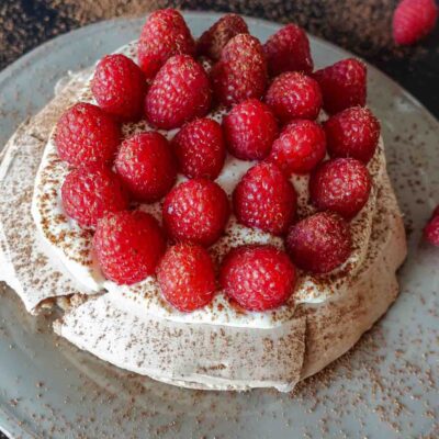 Raspberry pavlova on a black table with cocoa powder on a side.