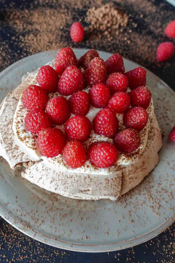 Raspberry pavlova on a black table with cocoa powder on a side.
