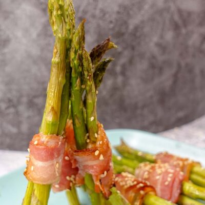 A close-up photo of six asparagus spears wrapped in bacon on a blue plate. The asparagus spears are trimmed and evenly spaced on the plate.