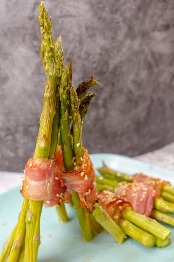 A close-up photo of six asparagus spears wrapped in bacon on a blue plate. The asparagus spears are trimmed and evenly spaced on the plate.