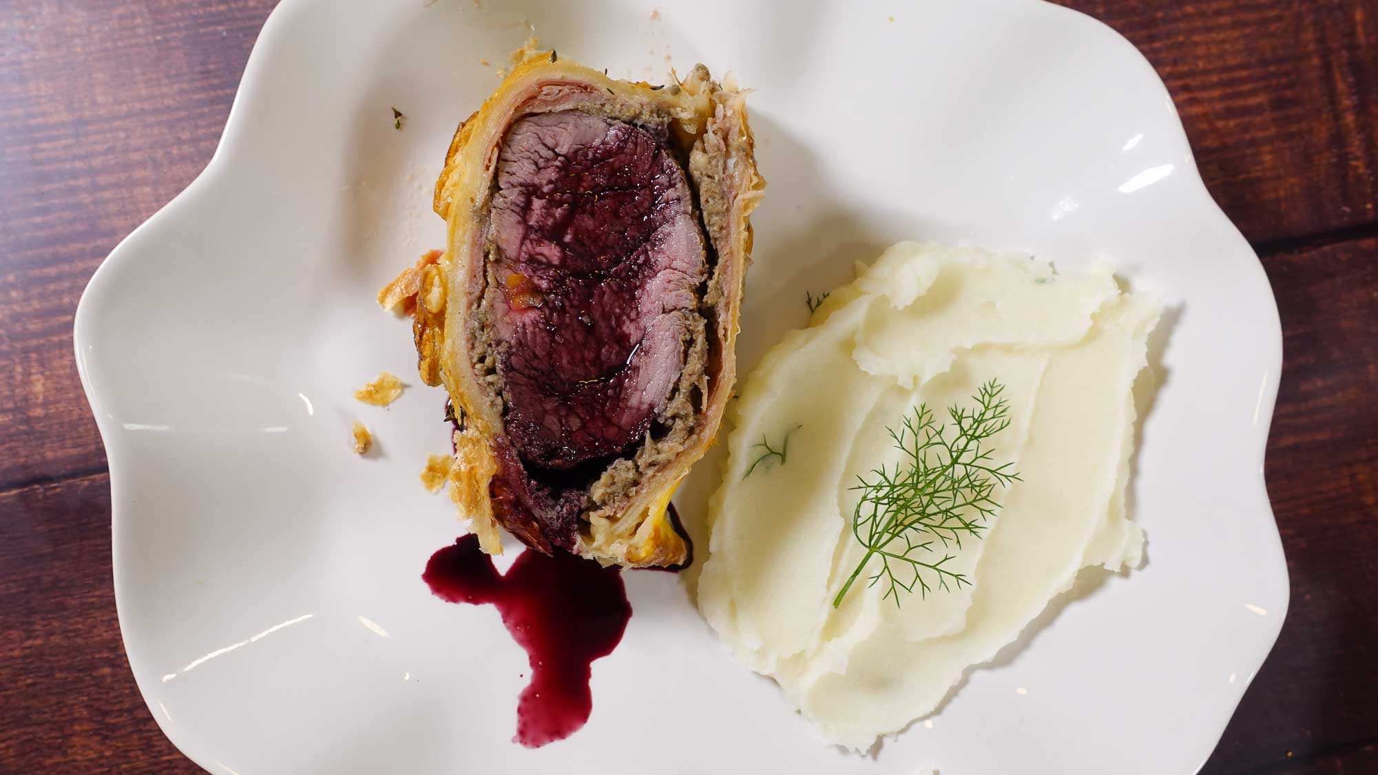 Cut Beef Wellington on a board with juicy meat inside and mashed potatoes and reduced wine sauce.
